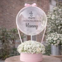 Naklejka na balon „I Komunia Święta” z symbolem krzyża na przezroczystym balonie bubble nad różowym flowerboxem z białymi kwiatami
