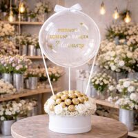 Naklejka na balon „Pierwsza Komunia Święta” w kolorze złotym na przezroczystym balonie bubble w flowerboxie z białymi kwiatami i bukietem z czekoladkami Rafaello