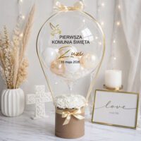Balon box komunijny ze złotą dekoracją, personalizowany balon z napisem „Pierwsza Komunia Święta Zuzia 10 maja 2026”, elegancka aranżacja z białymi kwiatami i świecą
